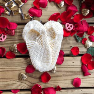 A pair of white slippers are laid out on a background of wooden slats, rose petals, chocolate hearts, and caramel candies.